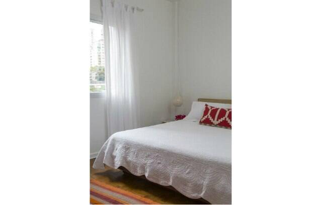 No quarto de casal, o branco é a cor principal. Almofadas vermelhas e tapete listrado criam ponto de cor no ambiente