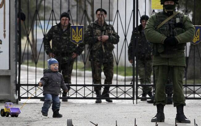Criança brinca perto de soldado russo (D) enquanto soldados ucranianos observam do outro lado do portão de base em Perevalne, Crimeia (4/3)