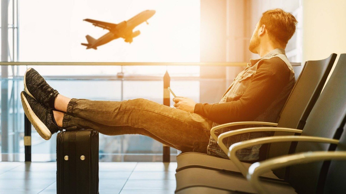 Homem com celular na mão e pés apoiados na mala, observando um avião decolar.