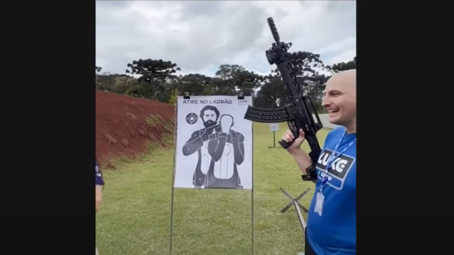 O empresário Luiz Henrique Crestani sorri com uma arma em mãos próximo a um alvo com a imagem do ex-presidente Lula com a frase 