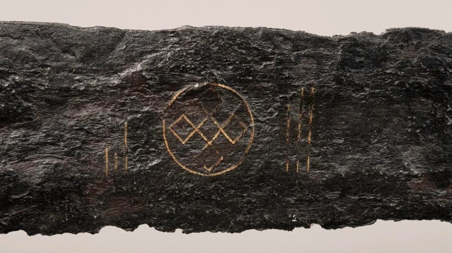 A lâmina exibe detalhes refinados em fio de liga de cobre, formando símbolos espirituais de grande valor ritual. De um lado, aparece uma roda solar — um círculo com uma cruz em seu interior — muito comum na Idade Média, principalmente na consagração de templos religiosos. A lâmina exibe detalhes refinados em fio de liga de cobre, formando símbolos espirituais de grande valor ritual. De um lado, aparece uma roda solar — um círculo com uma cruz em seu interior — muito comum na Idade Média, principalmente na consagração de templos religiosos.