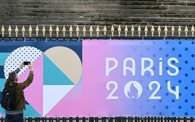 Homem tira foto de um cartaz dos Jogos Olímpico de 2024 em frente ao prédio da Assembleia Nacional Francesa, em Paris