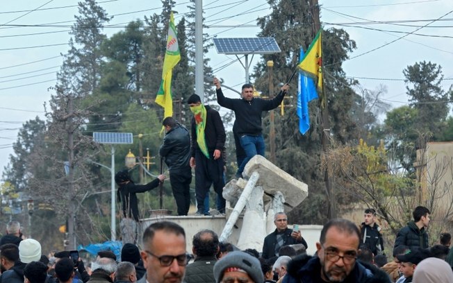 Curdos sírios celebram ao lado da estátua destruída do falecido presidente Hafez al-Assad, pai do atual presidente da Síria, Bashar al-Assad, enquanto comemoram a queda da capital, Damasco, para combatentes anti-governo, na cidade de Qamishli em 8 de dezembro de 2024