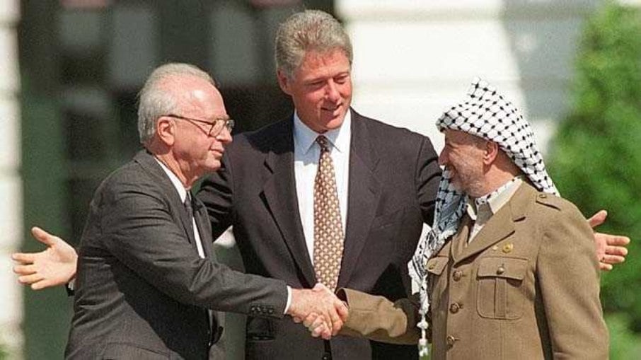 Yitzhak Rabin (à esquerda), aperta a mão de Yasser Arafat, observados pelo então presidente dos Estados Unidos, Bill Clinton.