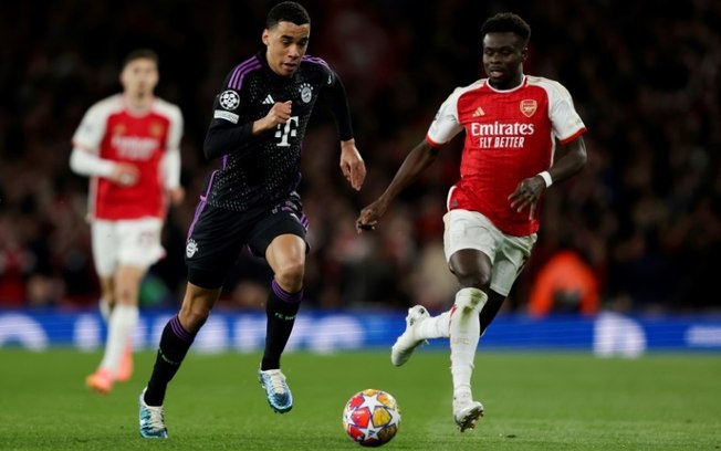 O atacante do Bayern de Munique, Jamal Musiala (à esq.), conduz a bola acompanhado pelo meia inglês do Arsenal, Bukayo Saka, durante o jogo de ida das quartas de final da Liga dos Campeões no Arsenal Stadium, em Londres, em 9 de abril de 2024.