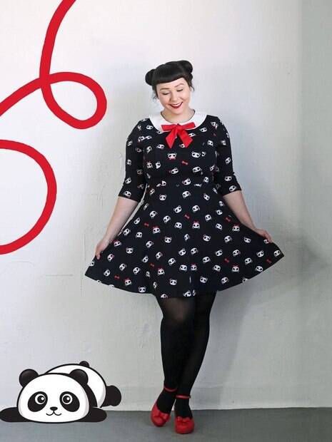 Mulher usando um vestido preto com pandas. Ela está de meia-calça preta e sapato vermelho. O fundo é branco e tem uma figurinha de panda próximo ao chão.