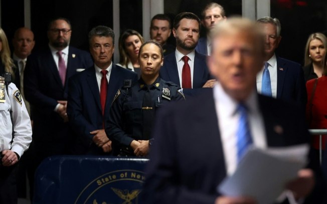 O senador JD Vance (ao centro, gravata vermelha), o senador Tommy Tuberville (atrás à direita, gravata azul) e Eric Trump (atrás ao centro) observa,  enquanto o ex-presidente dos EUA, Donald Trump, fala aos jornalistas durante seu julgamento em Nova York