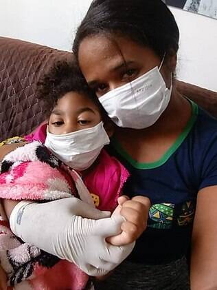 Na foto, Inabela Tavares (mãe) sentada no sofá com sua filha Gizella no colo, ambas usando máscara de proteção Mulher sentada no sofá com sua filha no colo, ambas usando máscara de proteção
