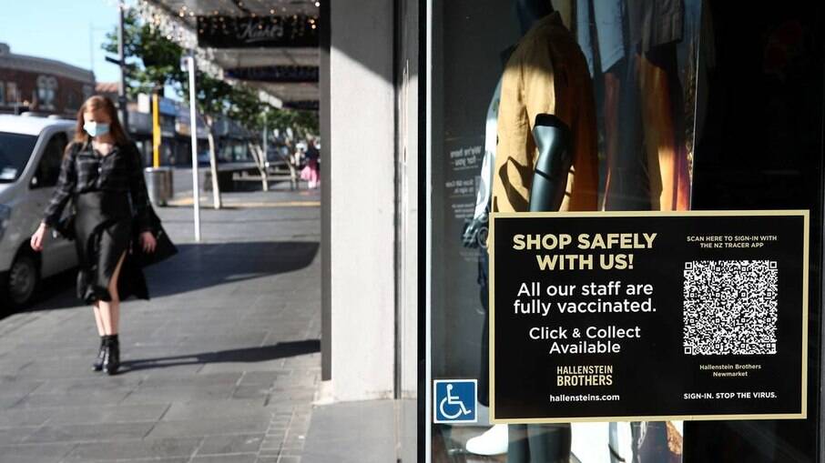 Mulher caminha em frente a loja que anuncia que todos os funcionários se vacinaram, em Auckland