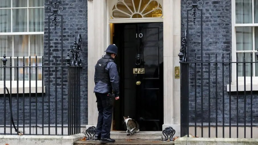 A imagem exibe a entrada de 10 Downing Street, a residência oficial e escritório do Primeiro-Ministro do Reino Unido em Londres. O policial de guarda está abrindo a porta para Larry, o gato