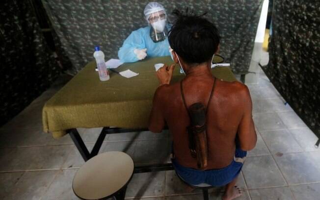 Em tenda de atendimento, homem indígena de costas é atendido, do outro lado da mesa, por profissional de saúde todo coberto por jaleco e face shield Em tenda de atendimento, homem indígena de costas é atendido, do outro lado da mesa, por profissional de saúde todo coberto por jaleco e face shield