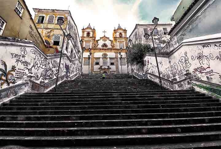 Escadaria da Igreja do Santíssimo Sacramento da Rua do Passo - Salvador (BA) - A escadaria conecta o bairro do Santo Antônio Além do Carmo ao Pelourinho e é famosa por sua arquitetura colonial e seu charme histórico. Composta por mais de 50 degraus de pedra, é cercada por casas coloridas e possui grande relevância no contexto cultural e religioso. Cenário de filmes e novelas, a escadaria evoca a essência do centro histórico de Salvador. Reprodução: Flipar