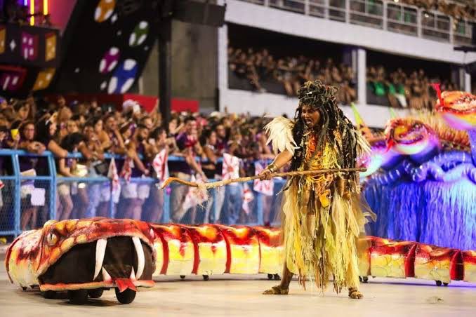 Uma grande serpente saiu do tripé que representava um ninho e deslizou pelo chão da avenida. Representando o ritual de preparação das guerreiras, uma mulher serpente foi a pivô nesse momento,com uma coreografia de tirar o fôlego. Reprodução/Twitter - 14.02.2024