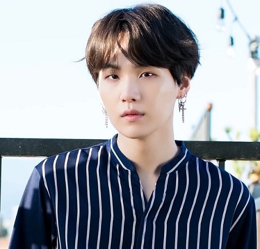 Suga, cujo nome verdadeiro é Min Yoongi, é um rapper, compositor e produtor do grupo BTS, o mais famoso de K-Pop no mundo. Ele é conhecido por suas letras introspectivas e por abordar temas como saúde mental e autoaceitação. Sua influência se estende além da música, impactando jovens em todo o mundo. Reprodução: Flipar